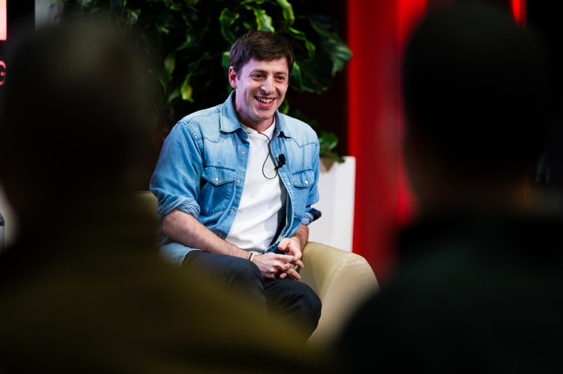 Alex Edelman sitting on stage wearing a white t-shirt underneath a chambray button down with a mic attached.