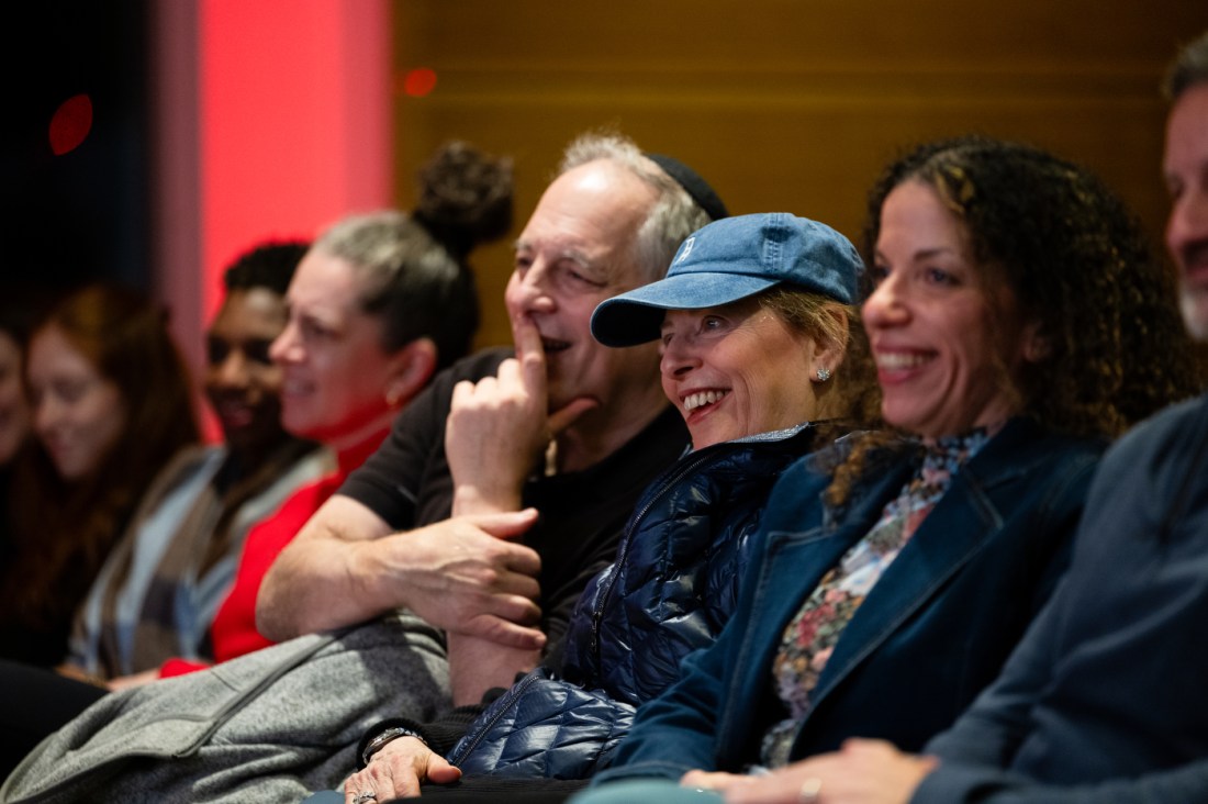 Close-up of engaged audience members laughing and enjoying the conversation. In focus is a woman wearing a blue baseball cap who is smiling broadly, with several other attendees visible beside and behind her.