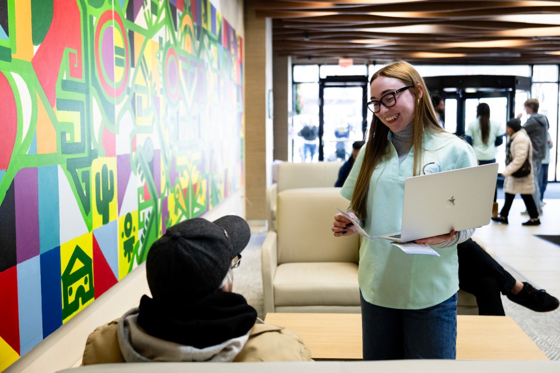 Mary Kathrine Hartigan standing in a hallway holding a laptop and smiling at a patient sitting on a bench. Mary is wearing a teal shirt over a grey turtleneck and glasses. The patient has on a jacket and a black baseball cap and is only visible from behind. 
