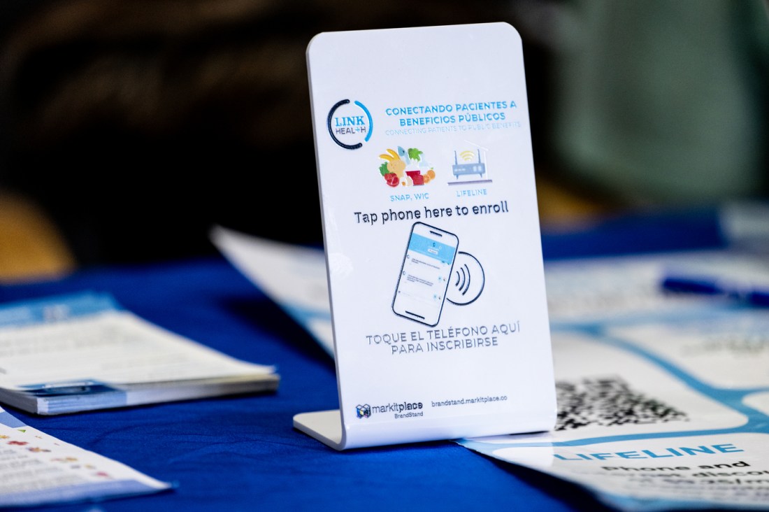 A white tabletop sign with tap-to-enroll NFC technology instructs patients to use their phone to register for SNAP, WIC, and Lifeline public benefits programs. 