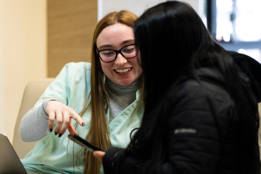 Mary Kathrine Hartigan sits next to a patient wearing a black jacket and points at their phone screen, explaining how to sign up for SNAP and WIC benefits. 