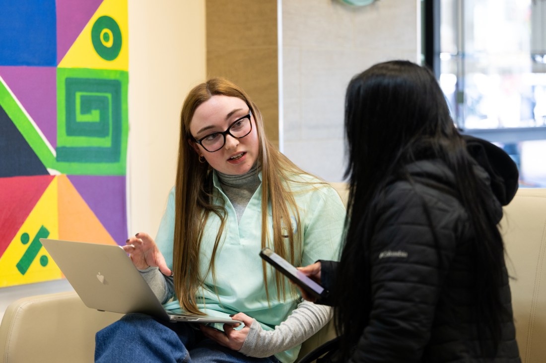 Mary Kathrine Hartigan sitting next to a patient in a black jacket. She is balancing a laptop in her lap and speaking tot he patient. She is wearing a teal shirt over a grey turtleneck and glasses. On the wall is a colorful art display of geometric shapes. 