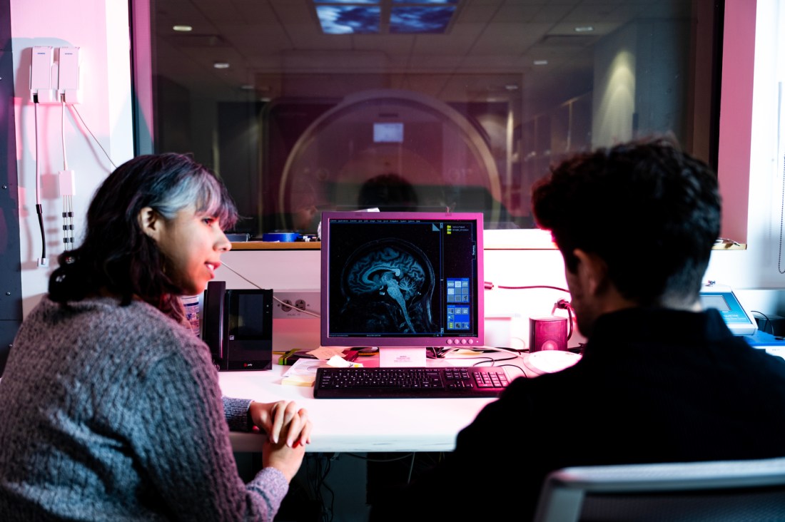 A woman and man sit at a computer discussing a brain scan taken on an MRI machine, visible through glass in another room.
