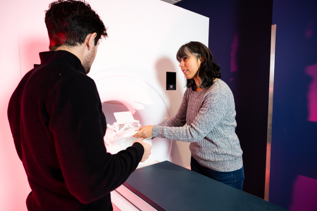 A man in a dark sweater stands on the left side of an MRI machine talking to a woman in a grey sweater on the right side of the machine.