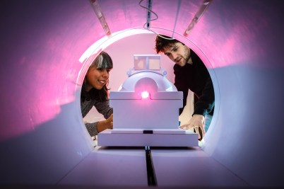 Two people look inside of an MRI machine. A pink light shines inside.