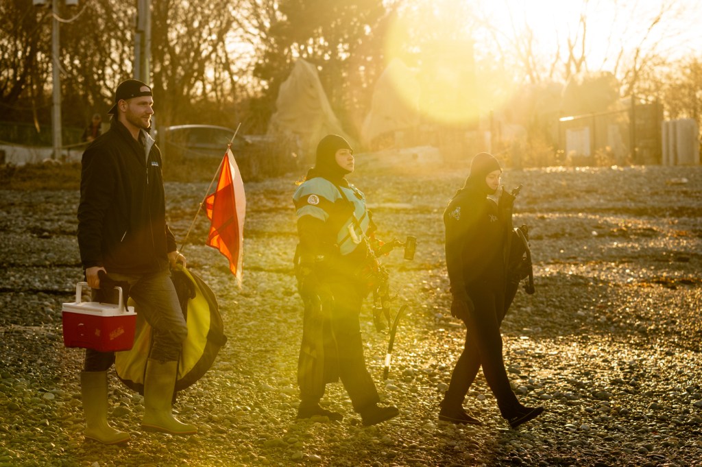 Three people walk across a rocky beach with sunlight shining across them. Two of them are dressed for snorkeling and the third carries a cooler.