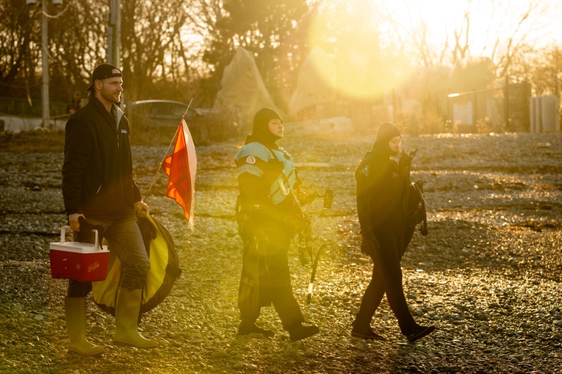 Three people walk across a rocky beach with sunlight shining across them. Two of them are dressed for snorkeling and the third carries a cooler.