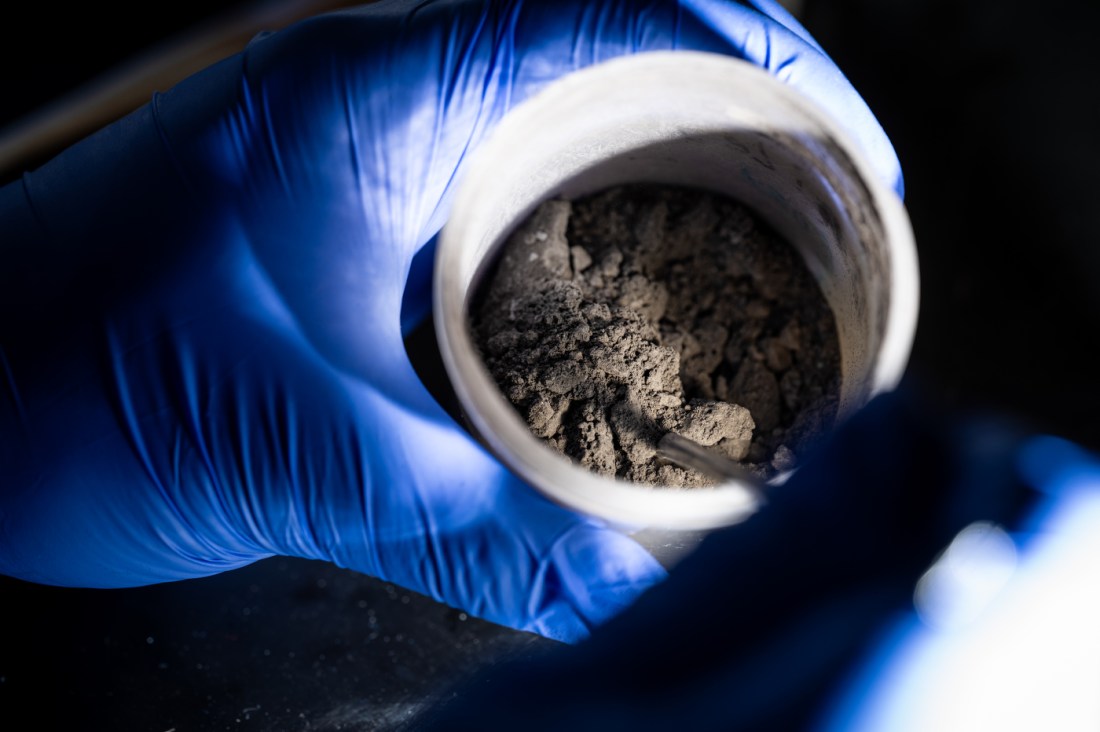 A hand wearing a latex glove measures out a sample of coal tailings from a jar held by another hand.