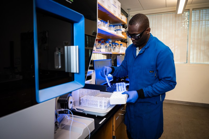 Lawrence Ajayi, wearing a lab coat, gloves and goggles, holds a tray of test tubes and removes one to inspect it.