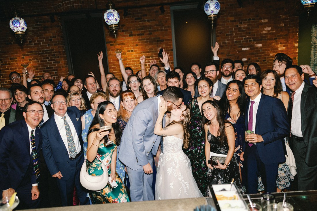 The newlyweds kiss while surrounded by a large group of wedding guests in formal attire, all gathered in front of an exposed brick wall decorated with blue mosaic hanging lights.