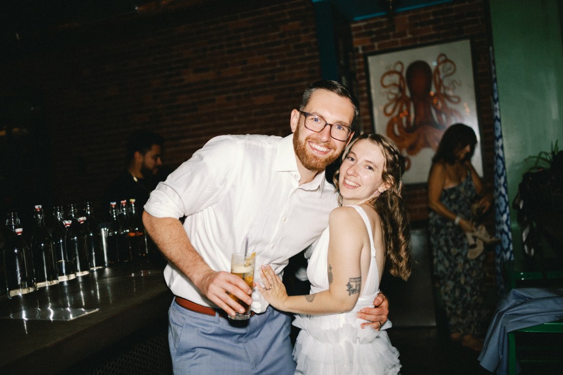 Ryan Kenny and Brenna Sorkin smile while embracing and holding drinks at an indoor party, with a brick wall, framed artwork, and other guests visible in the background.