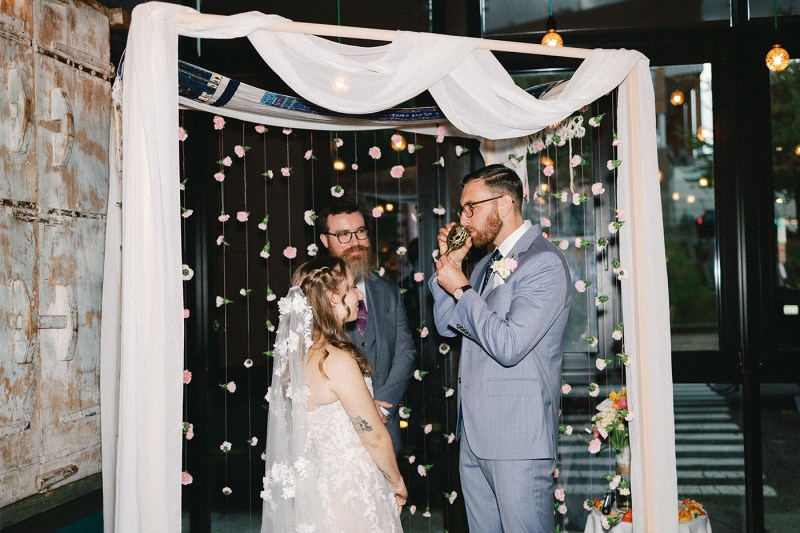 Ryan Kenny and Brenna Sorkin stand under a white draped archway decorated with hanging flowers and lights, with a black curtain backdrop during their wedding ceremony.