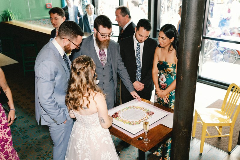 The couple signs their marriage document at a wooden table while surrounded by wedding guests, including the officiant and witnesses, in a venue with green walls and large windows.