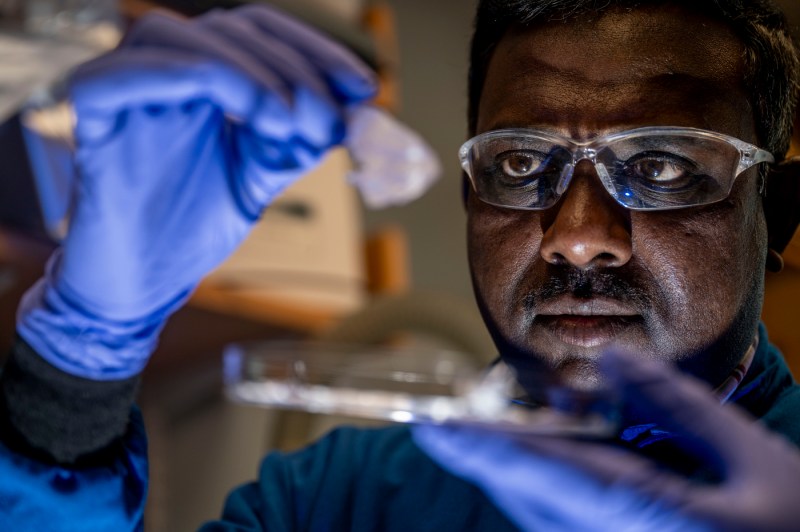 A close-up of a scientist who is wearing safety glasses and gloves holding up a piece of lab equipment. 