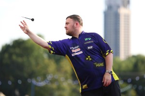 A man in a jersey displaying several sponsorships, throws a dart. The background features trees and a single tall building.