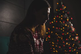 A person sitting in front of a Christmas tree in a dimly lit room looking sad.