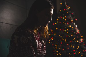 A person sitting in front of a Christmas tree in a dimly lit room looking sad.