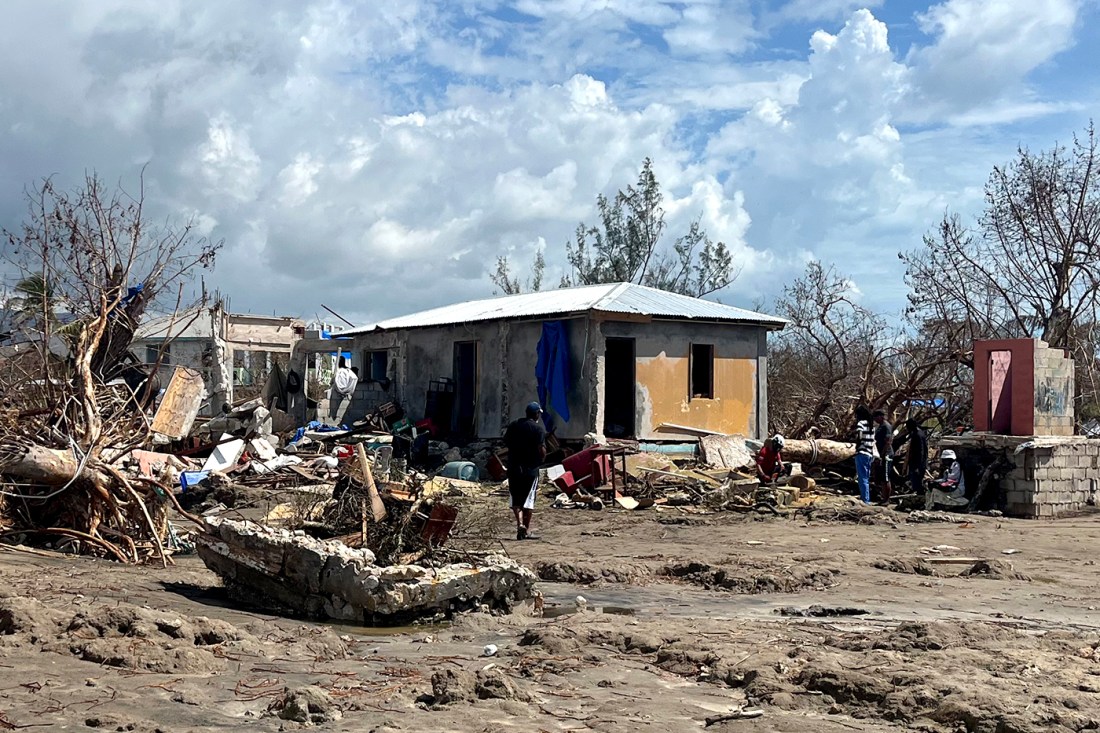 Several people linger around a one-story cement house missing windows and doors surrounded by debris and uprooted trees.
