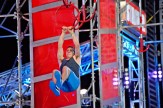 An athlete grips suspended rings mid-air on an obstacle course, wearing a dark sleeveless top and blue pants against a backdrop of dramatic red panels, metal scaffolding, and colorful stage lighting.