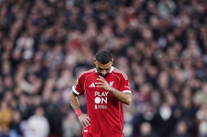 A professional footballer in a red jersey lowers his head on a field. The background is a blur of spectators.