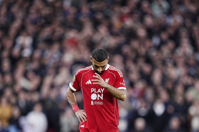 A professional footballer in a red jersey lowers his head on a field. The background is a blur of spectators.