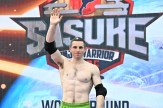 Josh Levin, shirtless and with short cropped hair, waves on the set of "Sasuke" with the silver text of the "Sasuke" logo behind him.