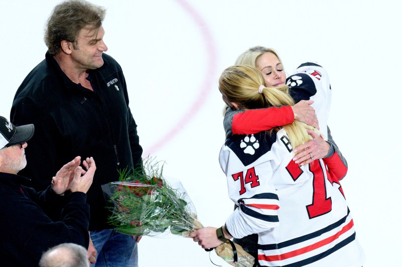 A Northeastern women’s hockey player in uniform hugs a woman on the ice during what appears to be a ceremony, with people holding flowers nearby.