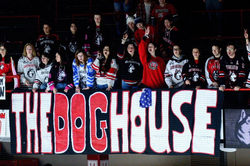 Northeastern hockey fans stand behind a large “THE DOGHOUSE” banner, cheering in the student section.