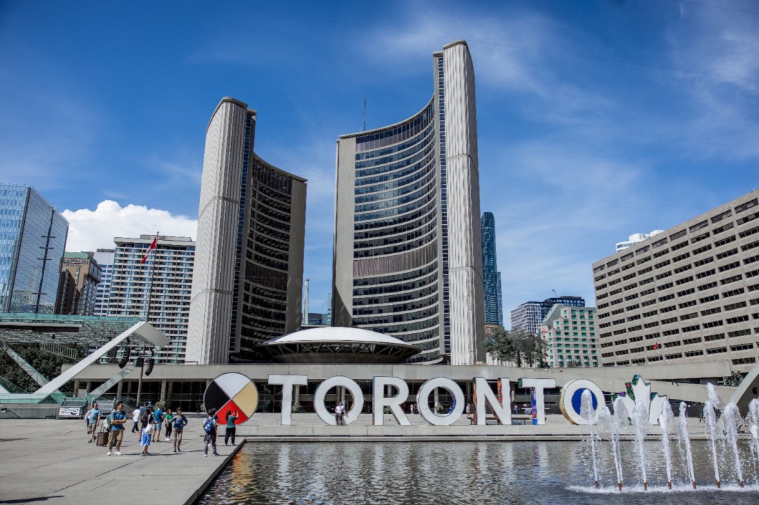 Concrete tall buildings surround a concrete square with a shallow pool of water with a fountain in the foreground and big letters spelling "Toronto" in the mid-ground along the horizontal edge of the pool. A group of people walks through the square along the vertical side of the pool.