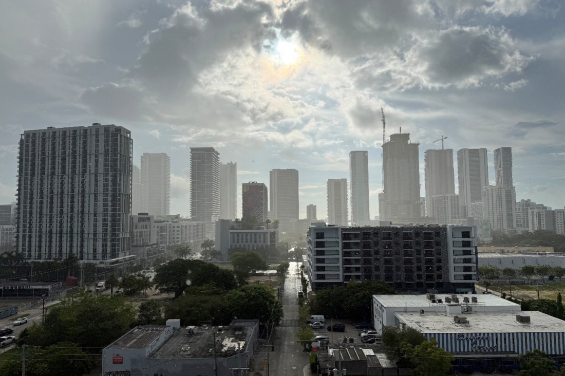 The morning sun breaks through the clouds and shines on concrete high-rises of Miami that look hazy against the sky. Lower buildings and green trees are seen in the foreground.