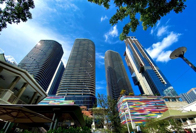 High-rises with colorful artwork at the bottom photographed from below against the blue sky and some green tree foliage.