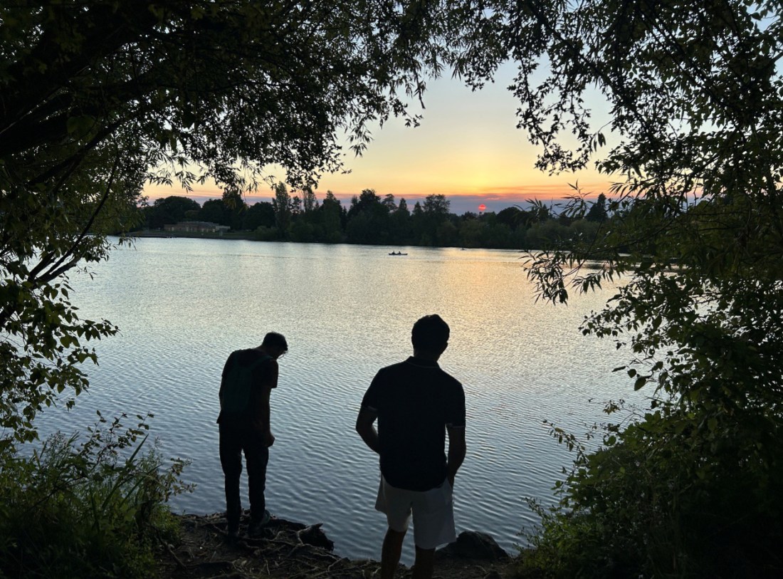 The photo shows two male silhouettes standing at the edge of a lake at sunset, framed by riparian vegetation. 