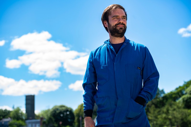 A man in a dark blue jacket stands against a light blue sky with just a few clouds.