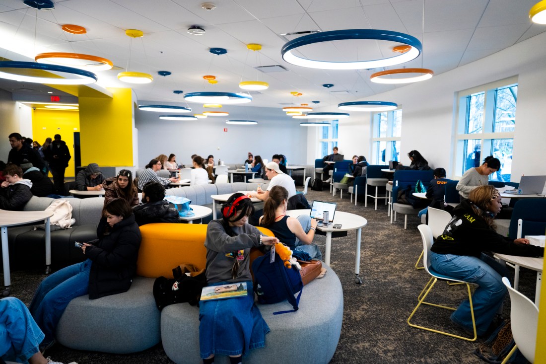 Groups of students study on laptops or scroll on their phones in a brightly lit study area.