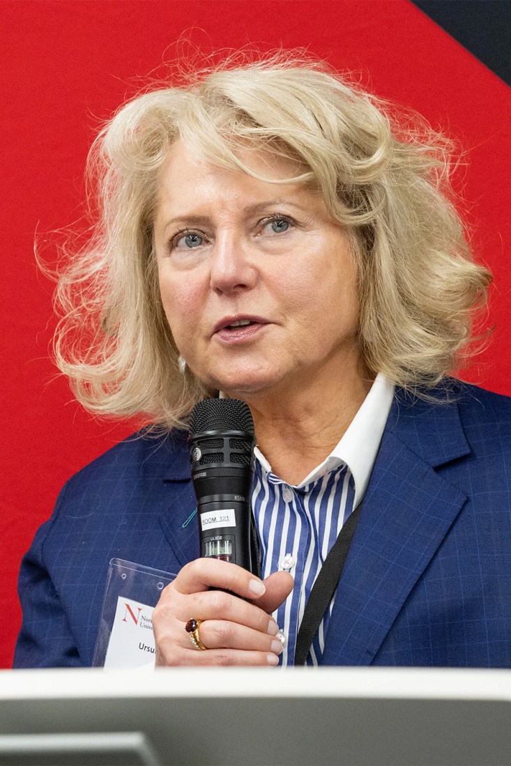 Ursula Smartt wearing a blue and white striped button down and a blue blazer speaking into a microphone. Smartt is sitting in front of a red background.
