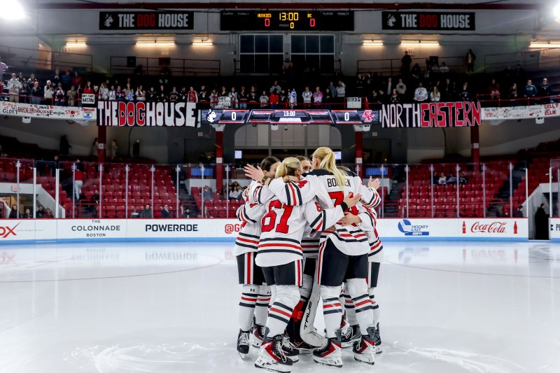 Members of the Northeastern women's hockey team huddle together on the ice.