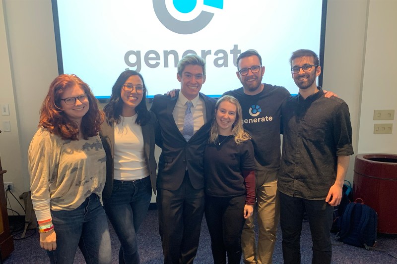 Six friends stand together smiling in front of a large Generate logo screen, with several wearing Generate-branded shirts, at an indoor event.