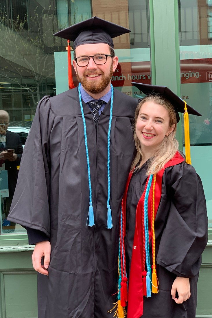 Ryan Kenny and Brenna Sorkin smile in graduation regalia, with Kenny wearing a blue honor cord and red tassel, and Sorkin wearing multicolored honor cords and a yellow tassel, standing in front of green glass walls.