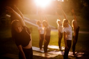 A group of women perform yoga in a park outdoors, each in the same pose, standing with their left arm extended overhead. The sun shine down from the top of the image.