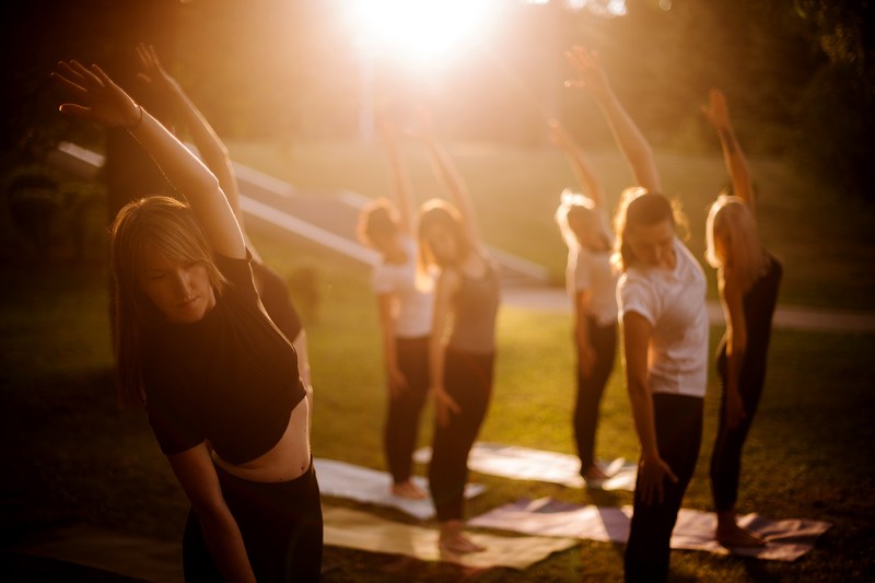 A group of women perform yoga in a park outdoors, each in the same pose, standing with their left arm extended overhead. The sun shine down from the top of the image.
