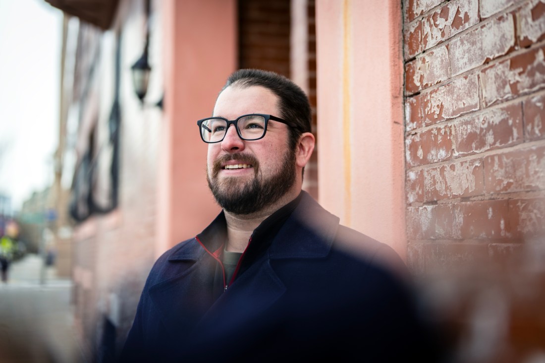 Portrait of Dan Nelligan wearing glasses and a blue quarter zip. Dan is standing in front of a brick building outside. 