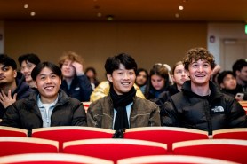 Three students sit together in a row smiling and waving at Blackman Auditorium.