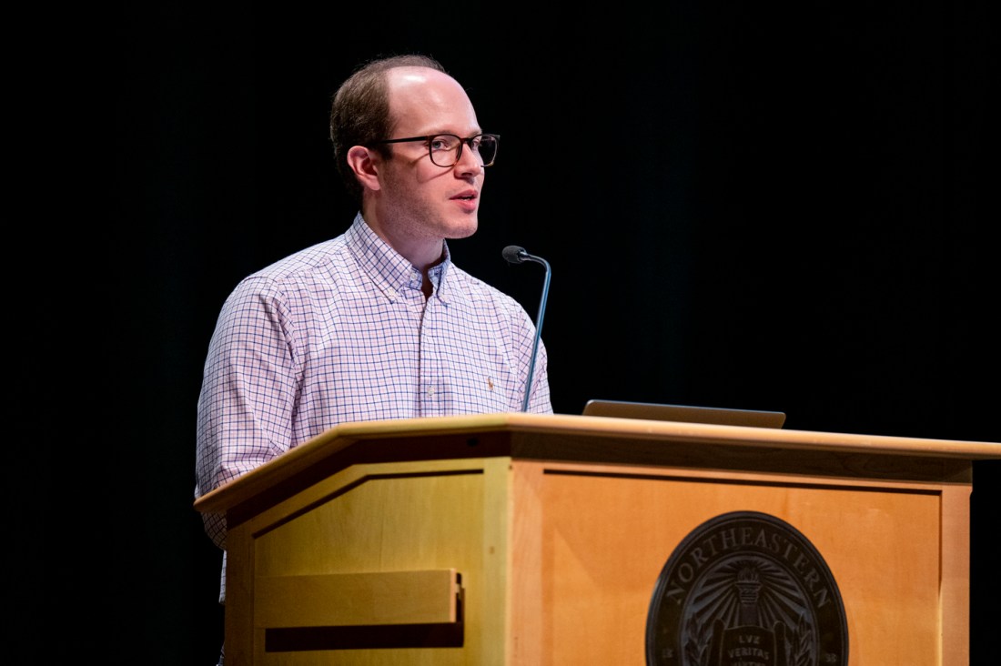 A person wearing a checkered button-down shirt and glasses stands at a podium speaking into a microphone.