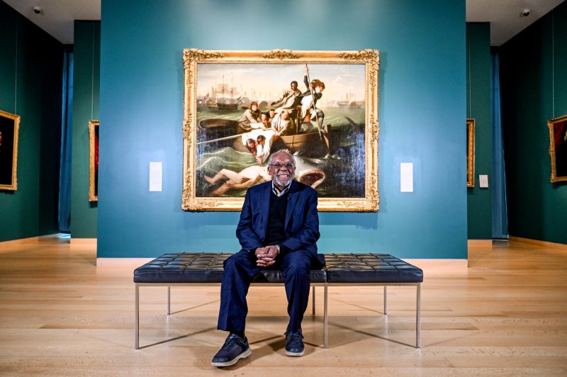 A man sits and poses for the camera with a large painting in the background.