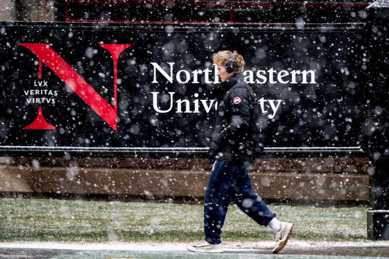 A students in a puffy jacket and headphones walks through snow with a "Northeastern University" sign in the background.