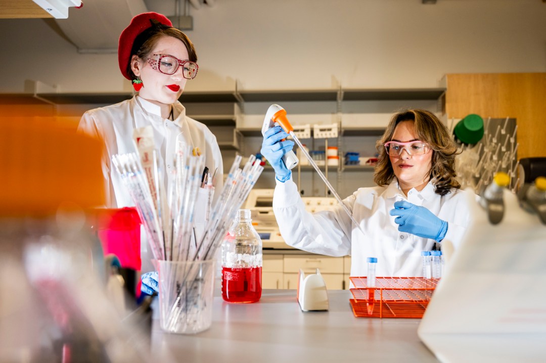 A white woman wearing a red berret, glasses, red lipstick and a white lab coat stands at the lab bench as another white woman with blond shoulder-length hair wearing a white lab coat and blue rubber gloves fills a glass test tube with a long dropper.