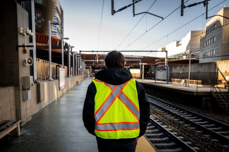 The back of a person a in reflective safety vest standing outside next to MBTA train tracks.