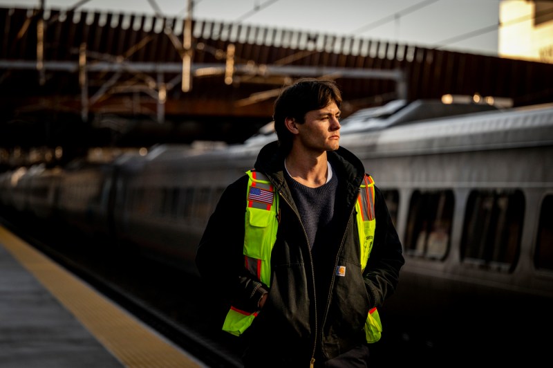A medium shot of a person in a reflective safety vest standing next to the MBTA train tracks.