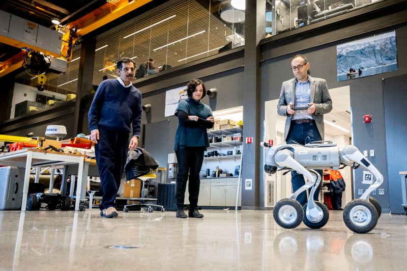 A low angle photo shows three people observing a display of a four-legged remote-controlled robot.