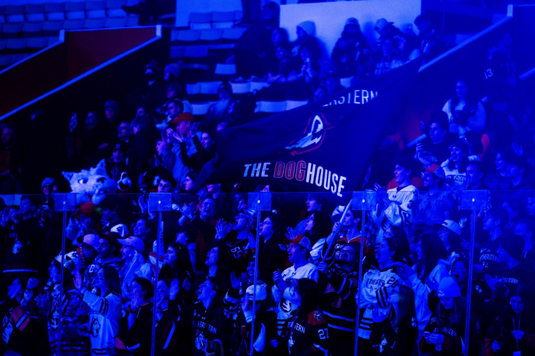Northeastern's student section is seen in the stands lit by dramatic blue lighting. Paws the Husky mascot stands off to the side with one paw pointed in the air and the fans are waving a giant black flag that says 'Northeastern The Dog House' on it.